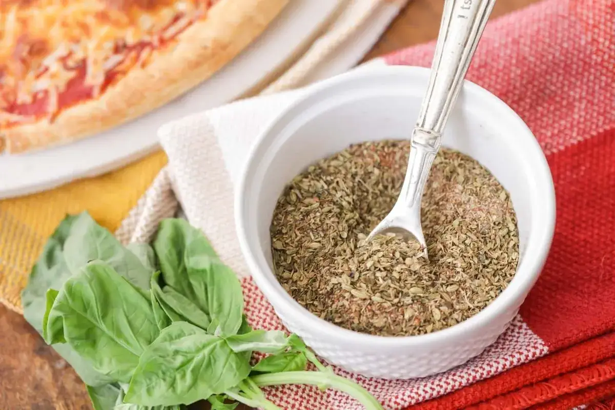 Mieszanka zi&oacute;ł, w tym oregano do pizzy, w białej miseczce z łyżeczką, obok świeżej bazylii i kawałka pizzy.