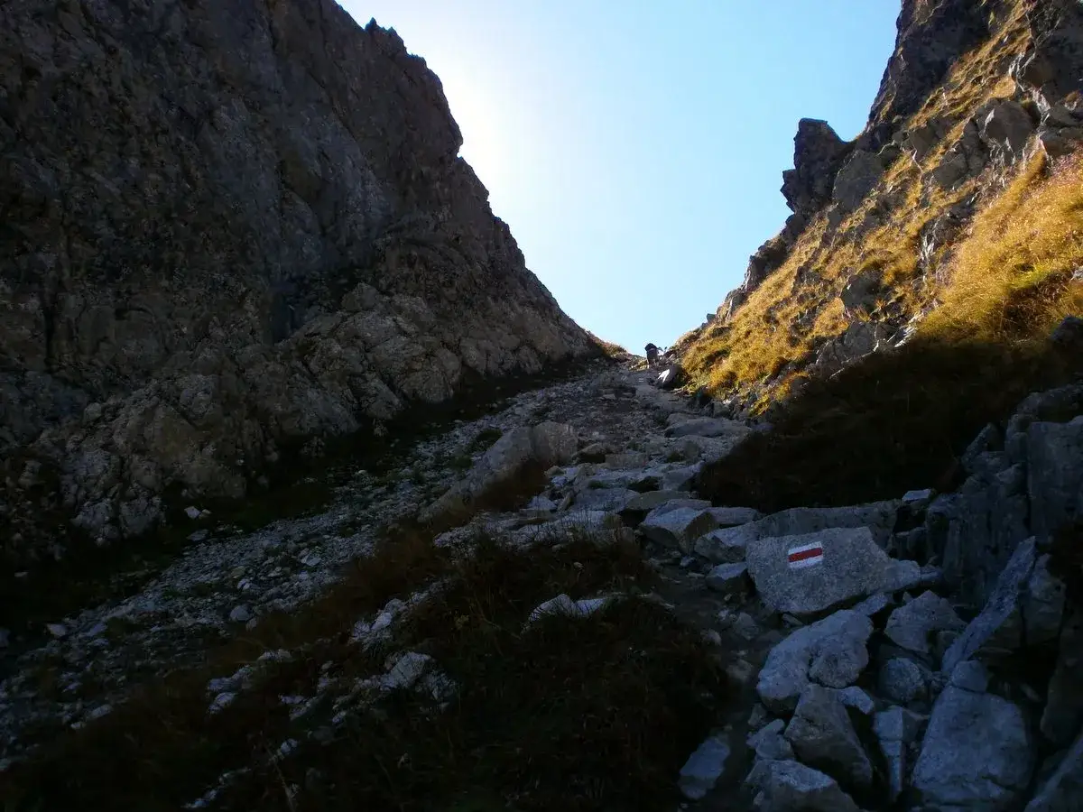 Kamienista ścieżka w g&oacute;rę, między skalnymi ścianami. W oddali widać turystę. To szlak przez Wrota Chałubińskiego.