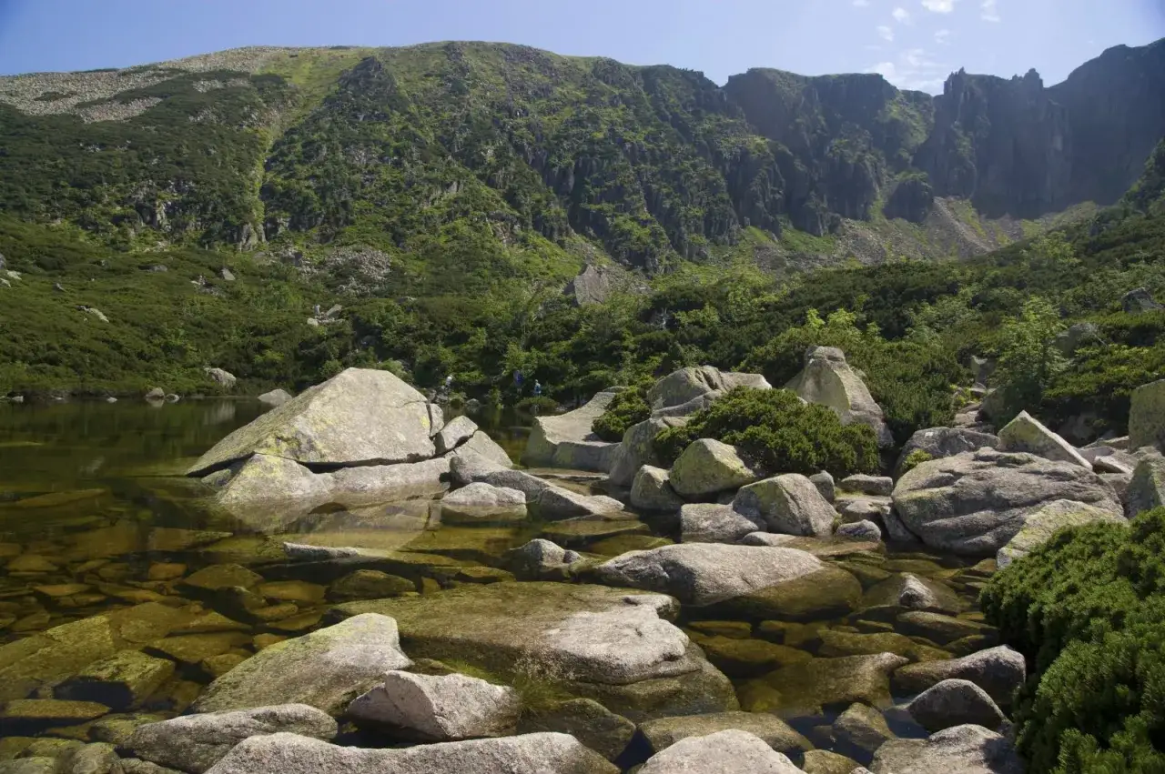 Kamieniste brzegi górskiego jeziora, gdzie woda odbija skaliste zbocza. W oddali widać turystów, a wokół rosną karłowate sosny. To malownicze śnieżne stawki.
