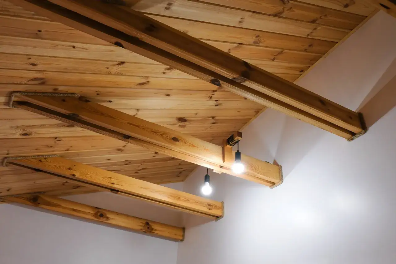 Drewniany sufit z belkami, oświetlony dwoma żar&oacute;wkami. Widać naturalne słoje drewna i sznurki owinięte wok&oacute;ł belek.