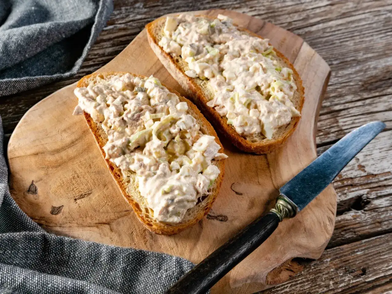 Dwa kawałki chleba z obficie rozsmarowaną pastą z tuńczyka, jajka i warzyw. Obok nóż i serwetka.