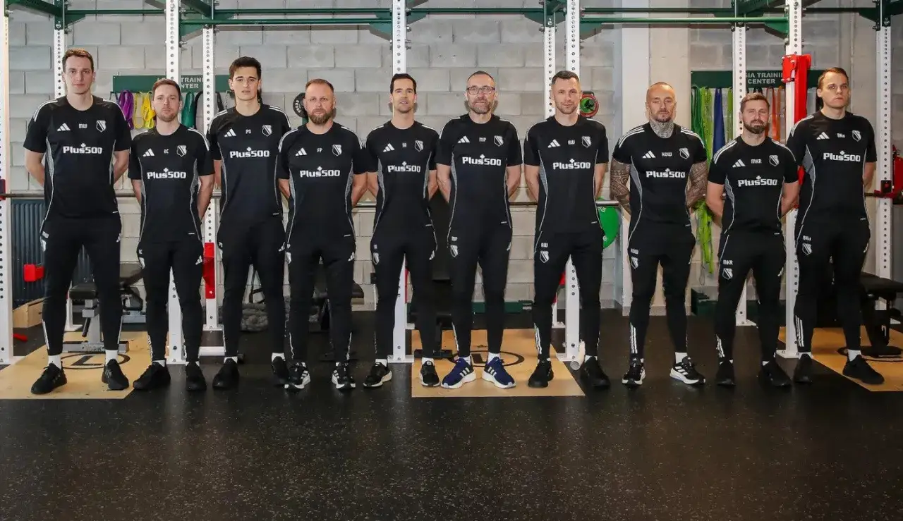 Grupa zawodników Legii Warszawa w czarnych strojach treningowych pozuje w siłowni.