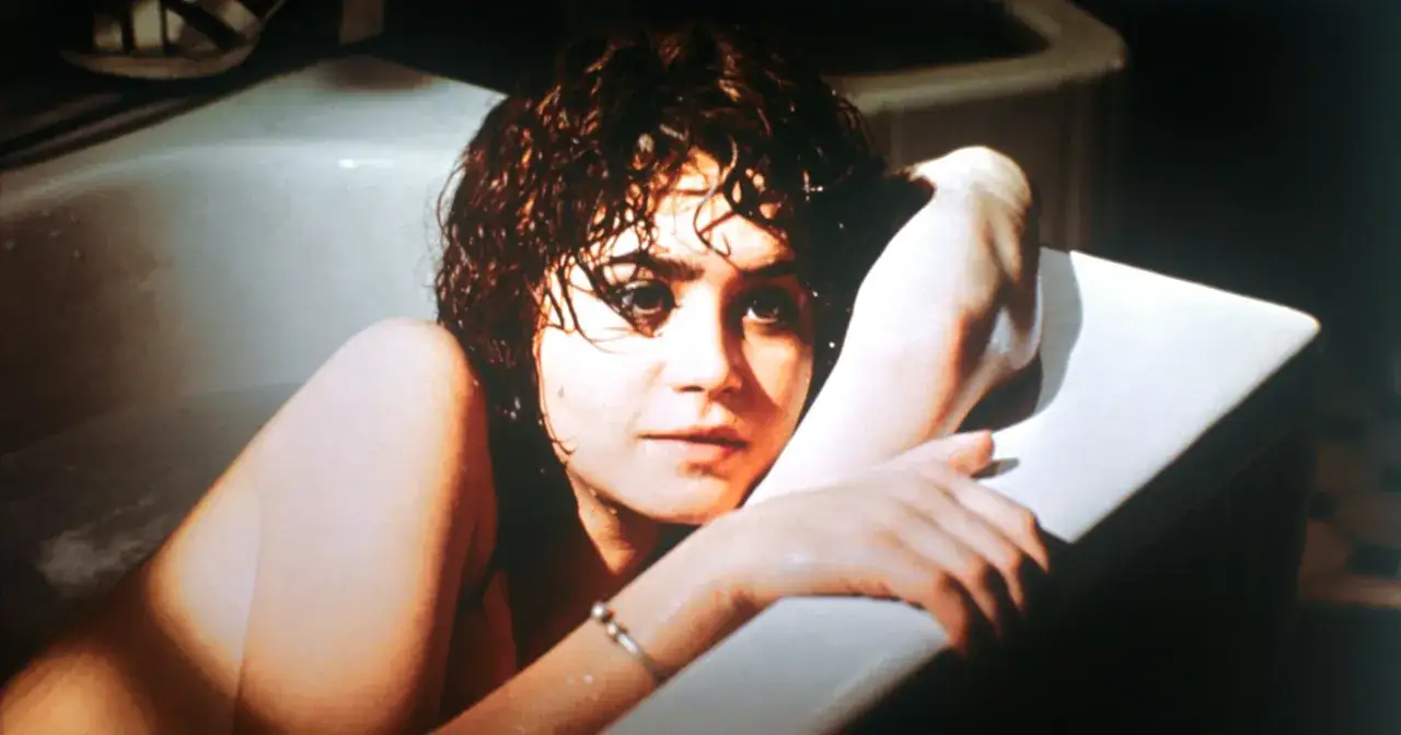 Młoda kobieta z mokrymi włosami leży w wannie, wspierając głowę na ramieniu. Scena przypomina 