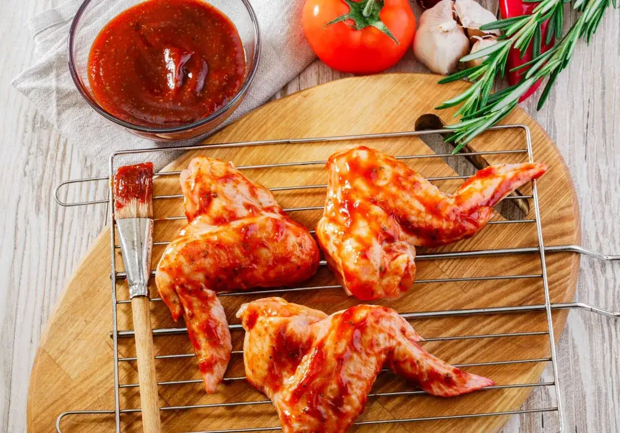 Skrzydełka kurczaka w marynacie na grilla, gotowe do pieczenia. Obok miseczka sosu i pędzelek.