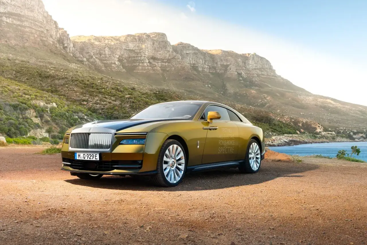 Złoty Rolls-Royce Spectre, elektryczny, testowany na tle skalistych gór i błękitnego morza.