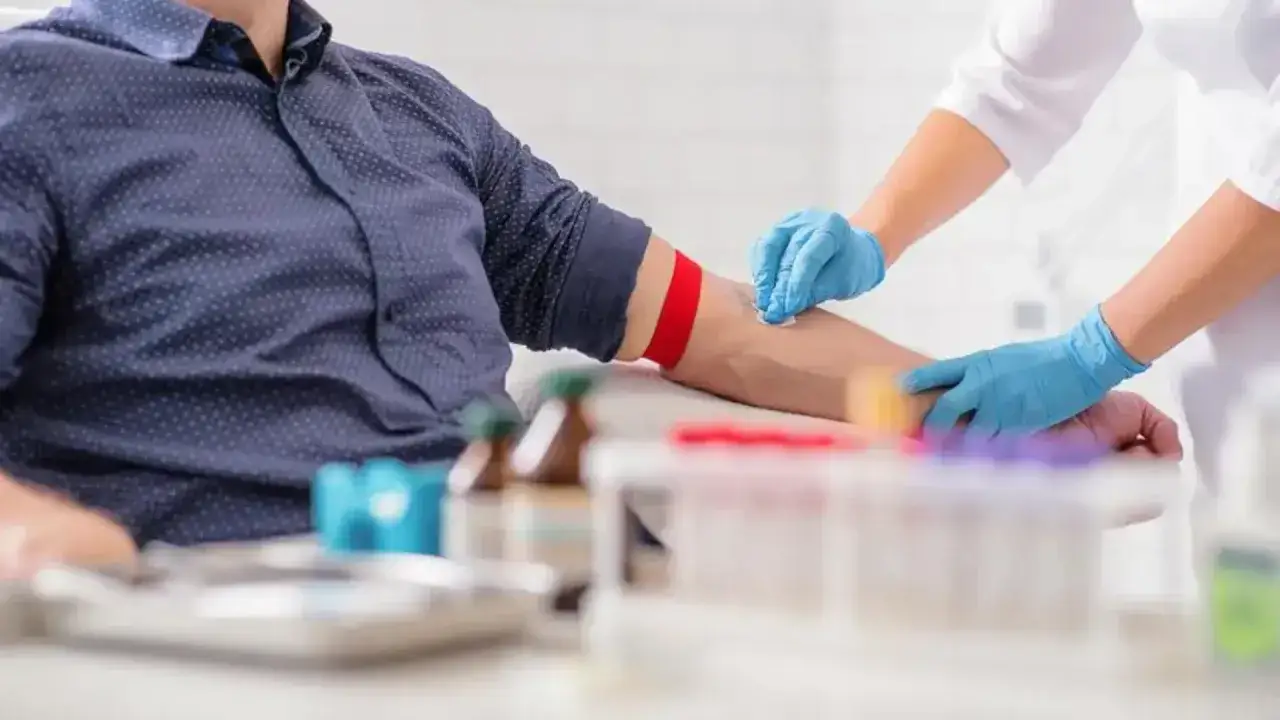 Opiekun medyczny w szpitalu pobiera krew od pacjenta. Ręka pacjenta jest zaciśnięta, a na ramieniu założony jest czerwony staz.