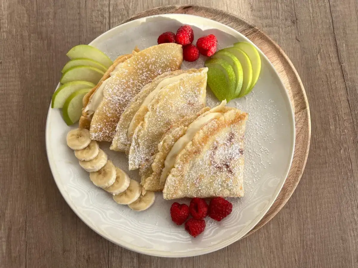 Pyszny przepis na zdrowe naleśniki z owocami: plasterki jabłka, banana i maliny zdobią talerz z naleśnikami posypanymi cukrem pudrem.