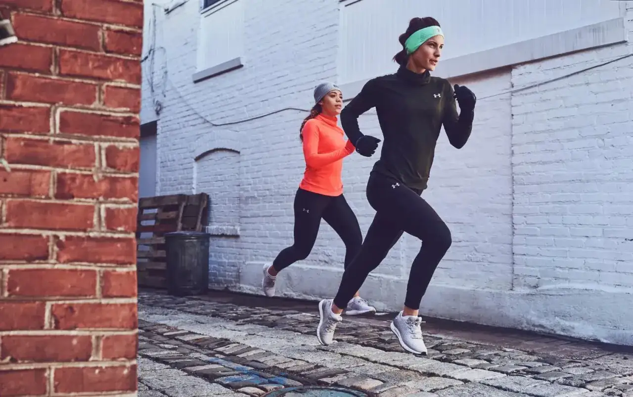 Dwie kobiety ćwiczą technikę biegu w miejskiej scenerii. Jedna w pomarańczowej bluzie, druga w ciemnozielonej.