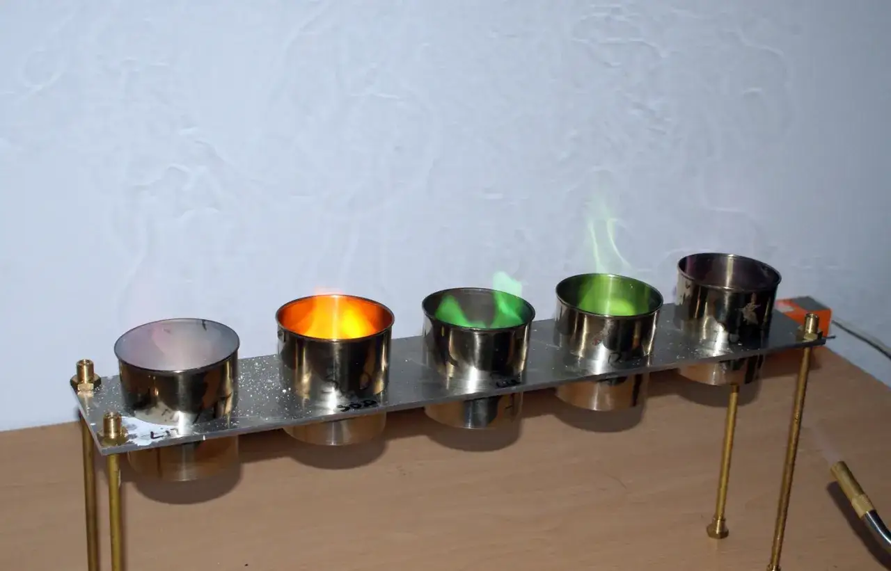 Eksperyment chemiczny: czy bimber się pali? Pięć metalowych naczyń z płomieniami – pomarańczowym i zielonym. Dym unosi się na tle ściany.