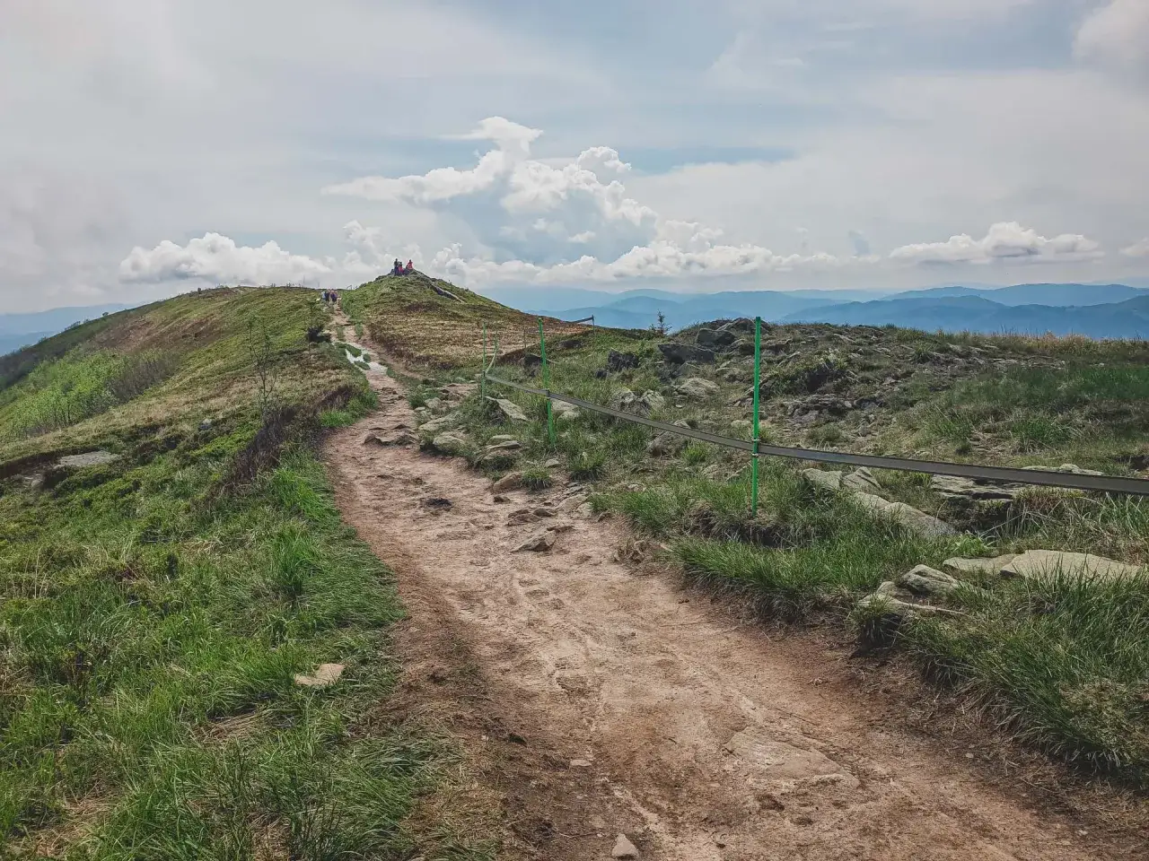 Kamienista ścieżka prowadzi na szczyt, gdzie widać grupę turyst&oacute;w. Rozległe, zielone połoniny Bieszczad&oacute;w rozciągają się aż po horyzont.