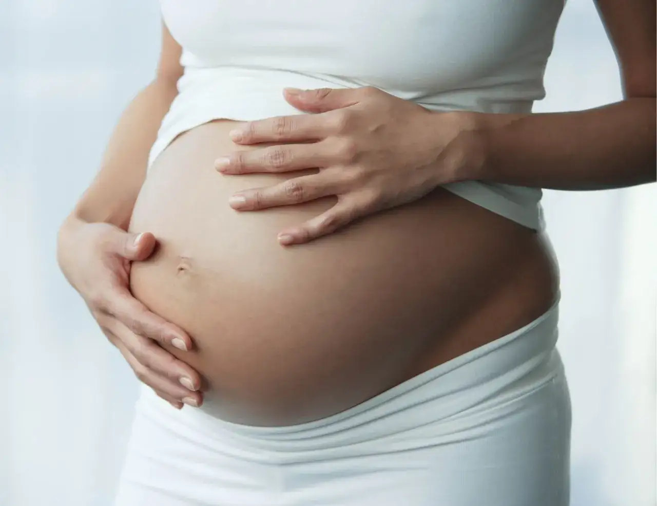 Brzuch kobiety w ciąży, delikatnie otulony dłońmi. Czy w ciąży można mieć orgazm? Tak, to normalne i zdrowe.