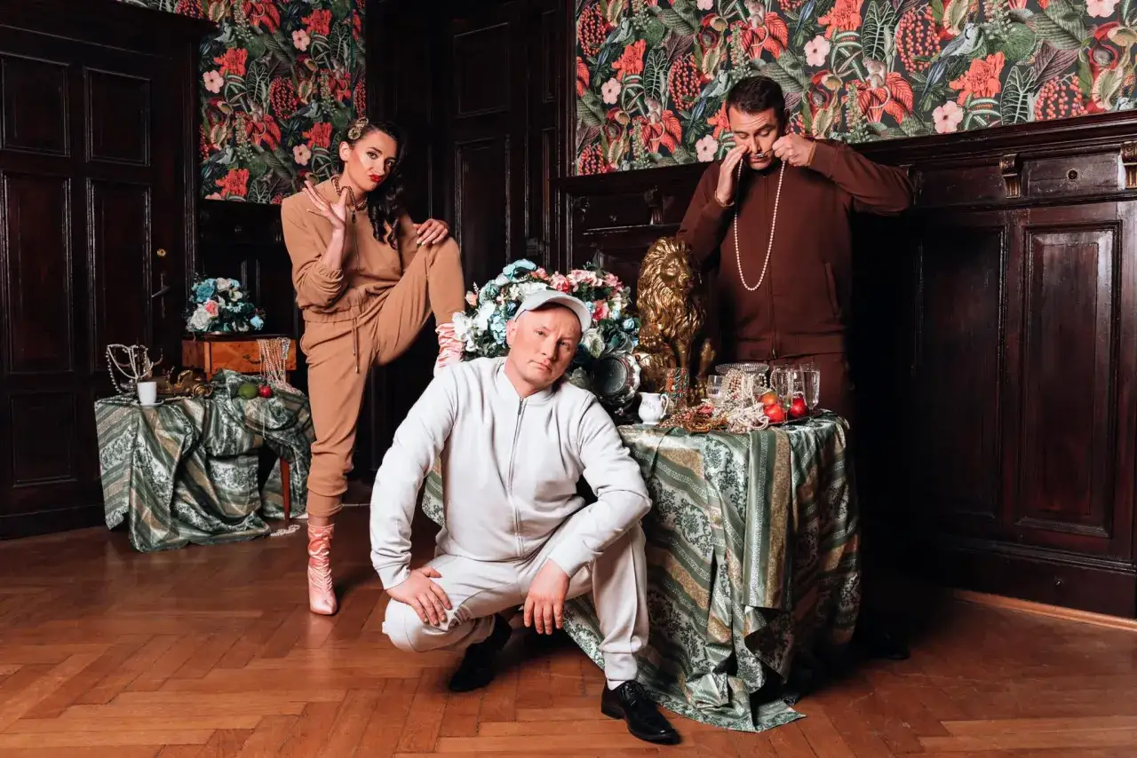 Trzy osoby w strojach sportowych pozują na tle tapety z motywem roślinnym. Jedna z nich, kobieta, gestykuluje. Obok stoi złoty lew. To scena z kabaretu K2.