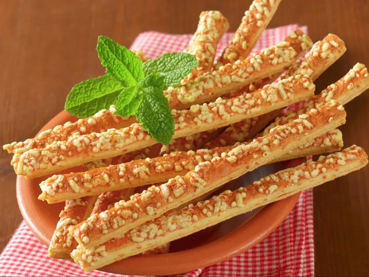 Chrupiące paluchy serowe z ciasta francuskiego, posypane startym serem, ozdobione gałązką mięty. Idealna przekąska na imprezę.