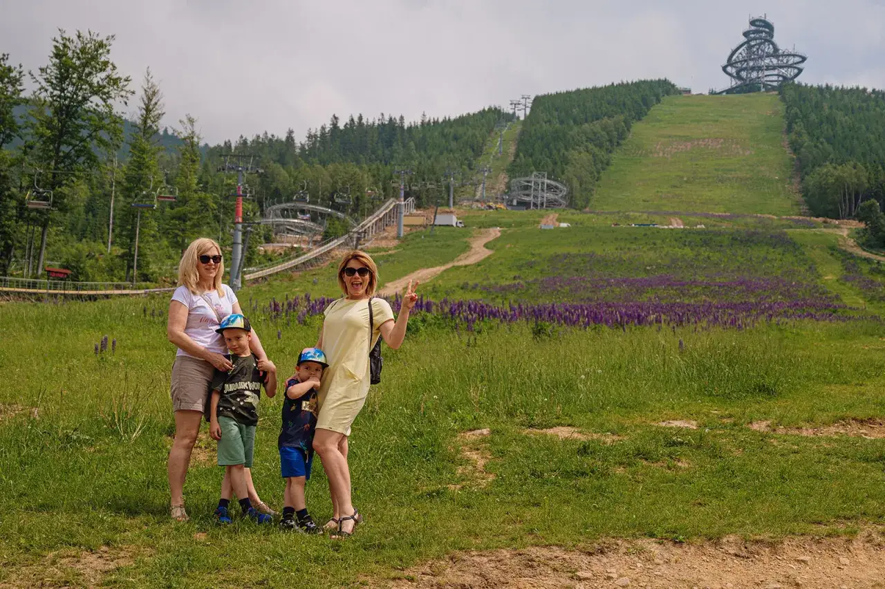 Rodzina na tle atrakcji Sky Walk w Czechach. W tle widać kolejkę linową i zjeżdżalnię.