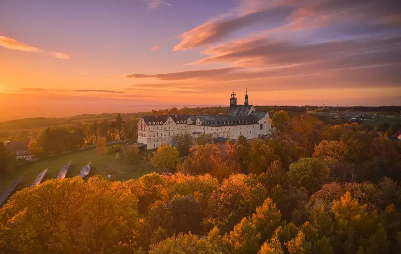 Zach&oacute;d słońca nad klasztorem na G&oacute;rze Świętej Anny. Jesienne drzewa otaczają zabytkowy budynek, tworząc malowniczy krajobraz.