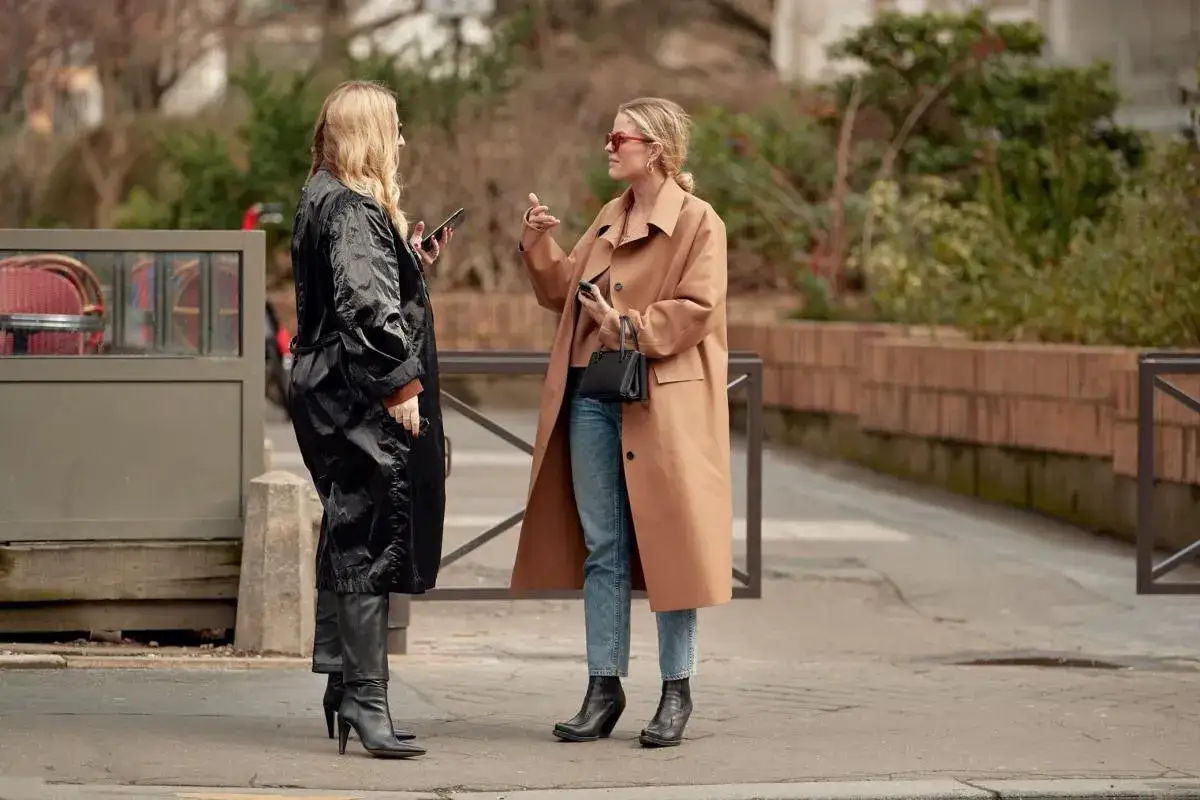 Dwie kobiety prezentują stylowe stylizacje na jesień. Jedna w czarnym płaszczu i kozakach, druga w beżowym płaszczu, jeansach i botkach.