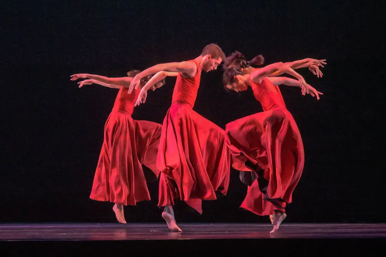 Trzech tancerzy w czerwonych sukniach na scenie, uchwyceni w dynamicznym ruchu. Ich gesty i pozy oddają dramatyzm i piękno 