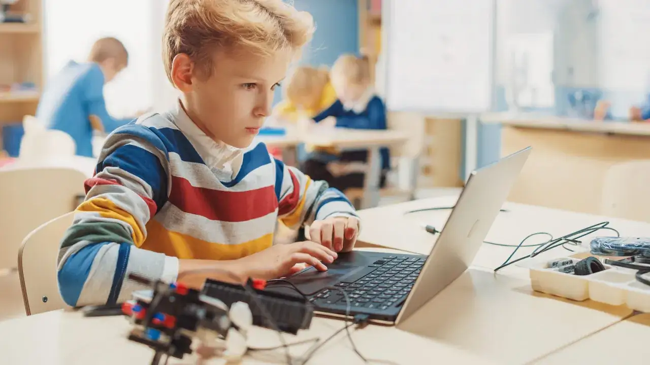 Chłopiec w kolorowym swetrze uczy się informatyki klasa 1, pisząc na laptopie. Obok leżą kable i części do robotyki.