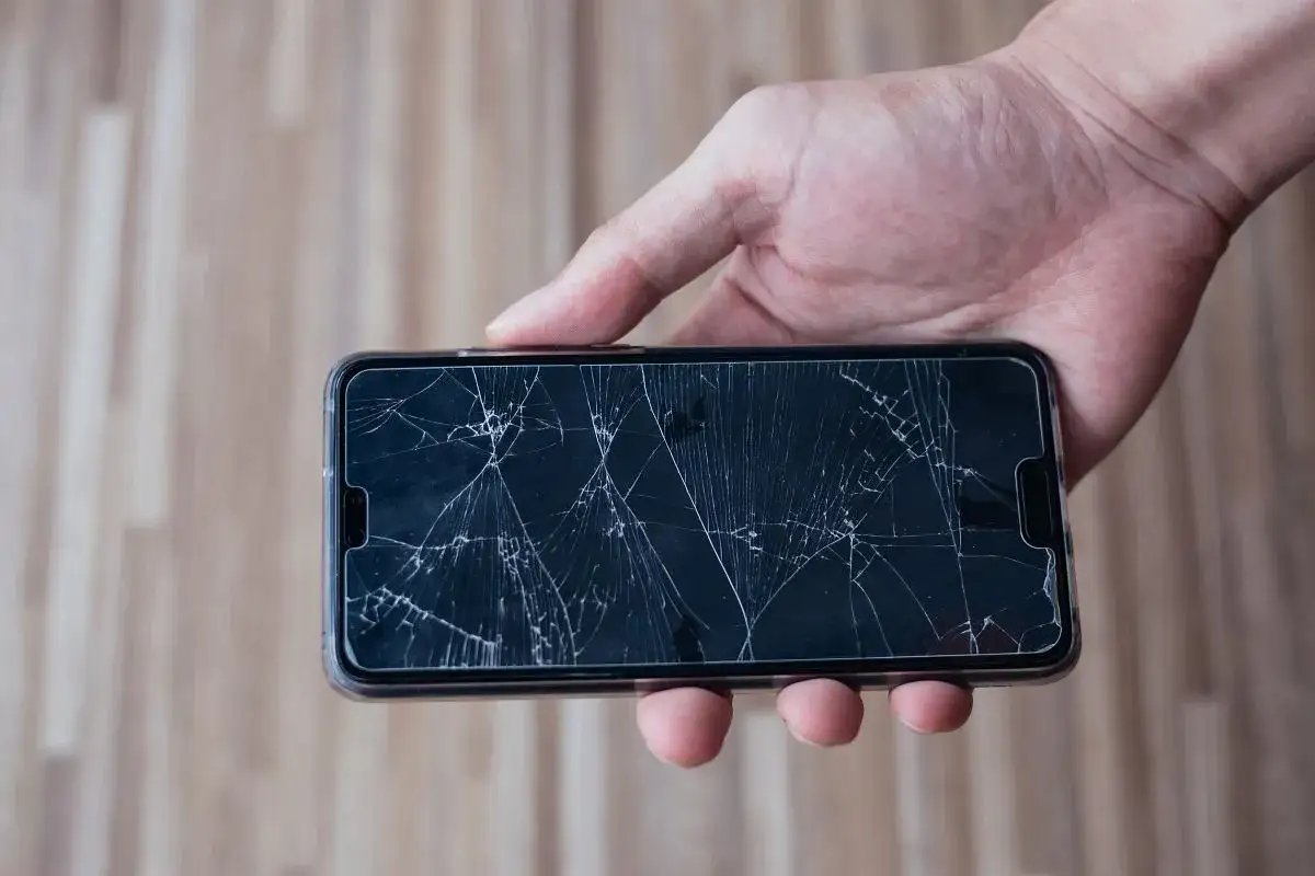 Dłoń trzyma telefon z mocno pękniętą szybką. Czy można używać telefonu z pękniętą szybką? To pytanie zadaje sobie wiele os&oacute;b.