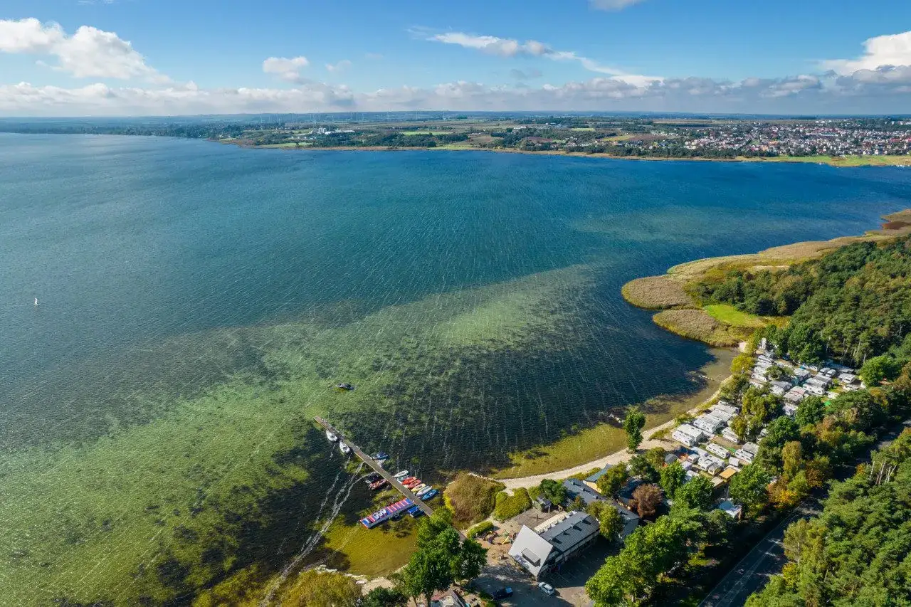 Malowniczy widok na jezioro i plażę na cyplu helskim. Widać łodzie przy pomoście, kemping i las.