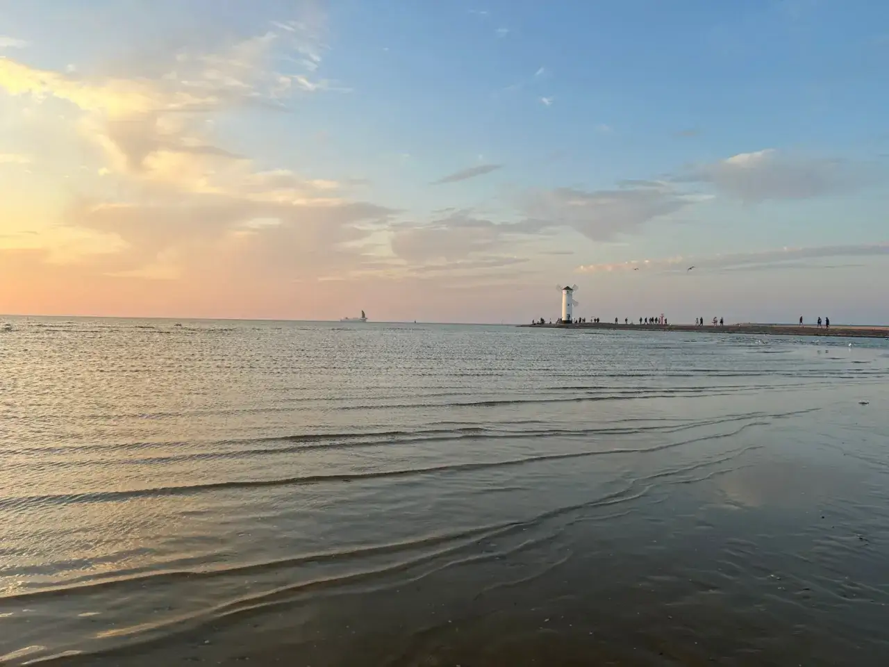 Zachód słońca nad morzem. Na horyzoncie biała latarnia morska i spacerujący ludzie. Aktualny stan morza spokojny, z delikatnymi falami.