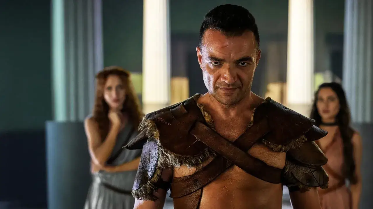 Spartakus, serial gdzie obejrzec? Gladiator w zbroi, zdeterminowany, z kobietami w tle.