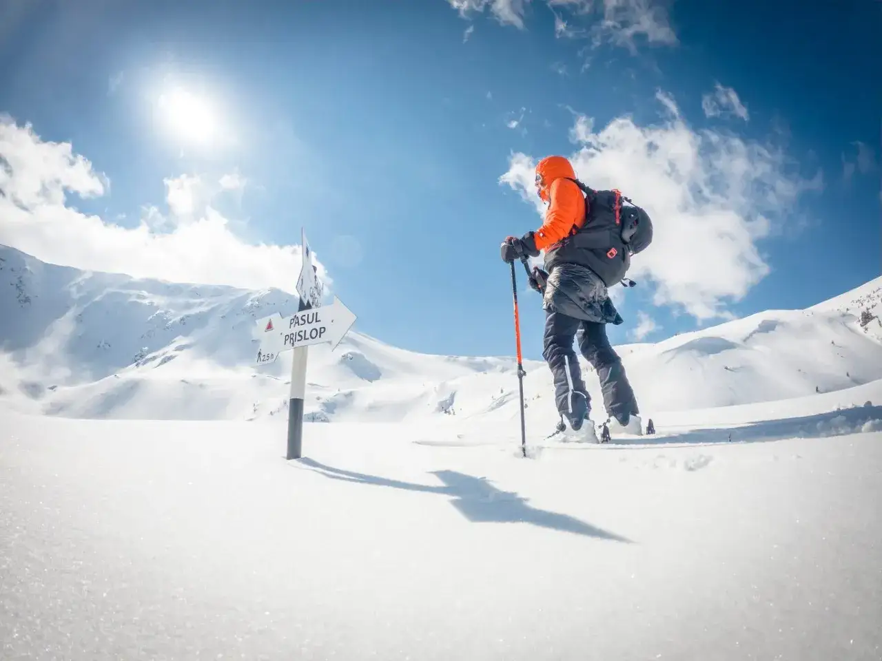 Osoba na nartach skiturowych podąża za znakiem wskazującym drogę na Przełęcz. Słońce świeci na zaśnieżone góry.