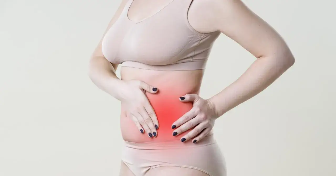 Kobieta trzyma się za brzuch, gdzie widoczny jest czerwony obszar bólu, sugerujący problemy z narządami po lewej stronie brzucha.