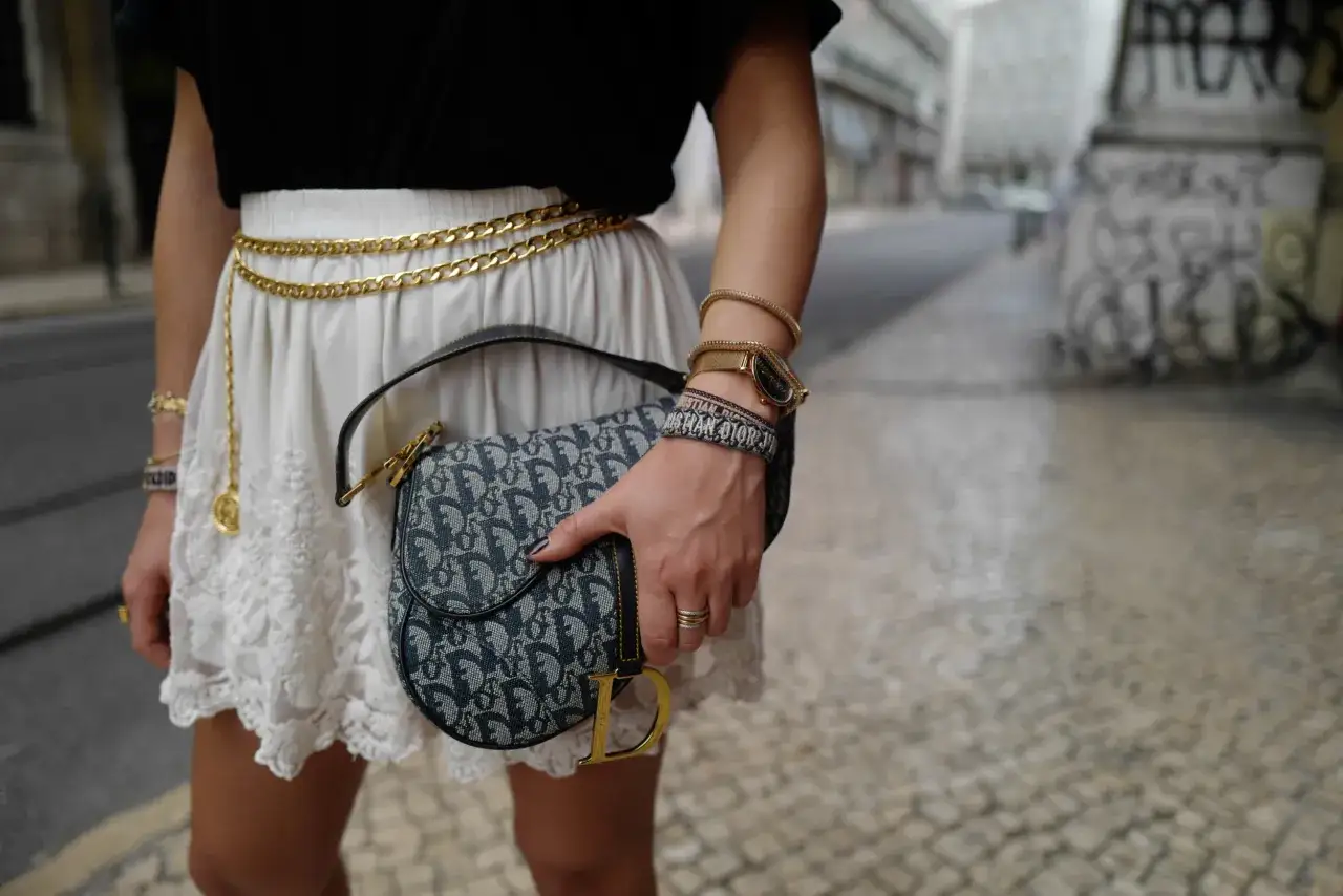 Elegancka kobieta z torebką Dior Saddle na tle miejskiej ulicy. Złoty pasek i biżuteria dodają blasku.