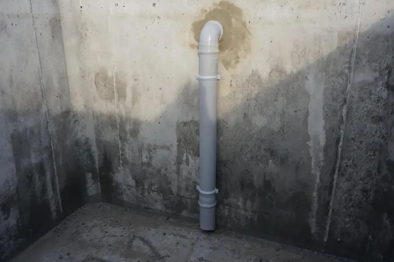 Szara rura do szamba w betonowym narożniku, zamocowana do ściany.