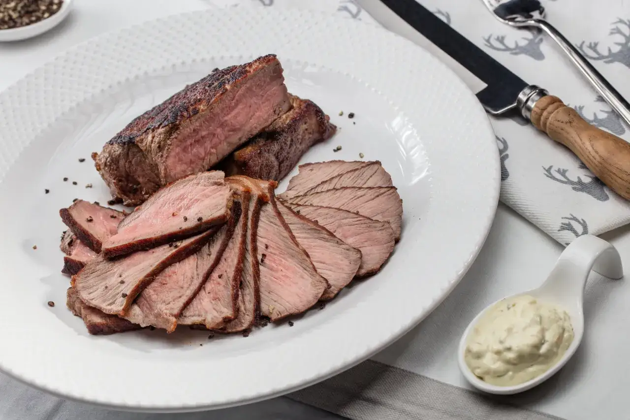 Soczysty stek z rostbefu, idealnie wysmażony, pokrojony w plastry i posypany pieprzem. Obok sos i sztućce.