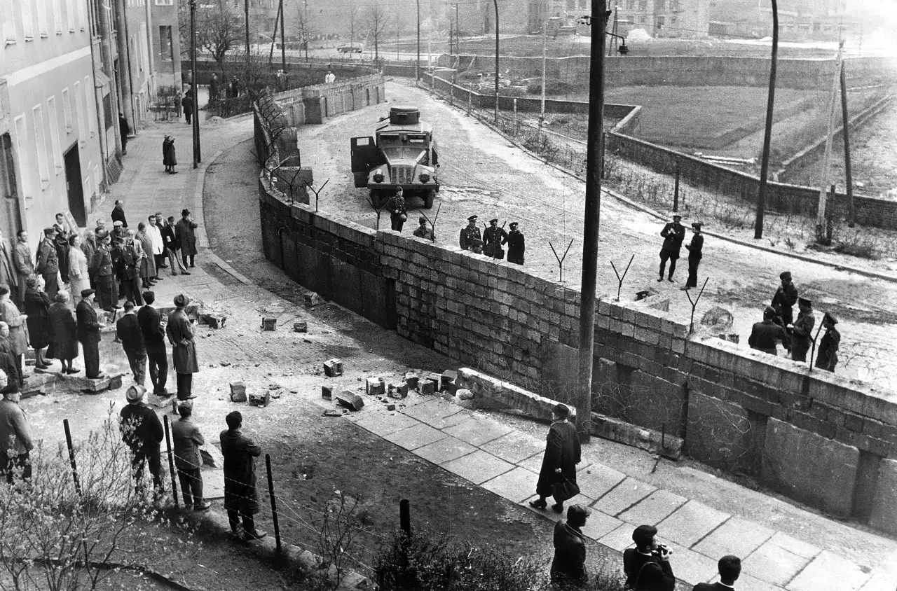 Ludzie obserwują budowę muru berlińskiego, żołnierze pilnują terenu, a na drodze stoi wojskowy pojazd.
