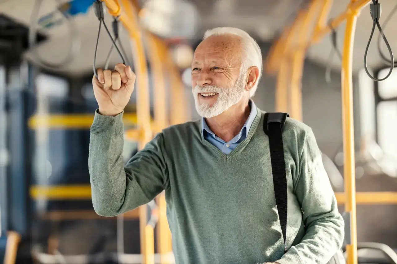Uśmiechnięty starszy pan z siwą brodą trzyma się poręczy w autobusie. Zastanawia się, jak długo można pracować po osiągnięciu wieku emerytalnego.