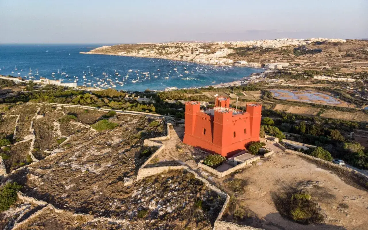 Czerwona wieża na wzgórzu, widok na zatokę pełną łodzi i miasto. Malta co zobaczyć - idealne miejsce na wycieczkę.
