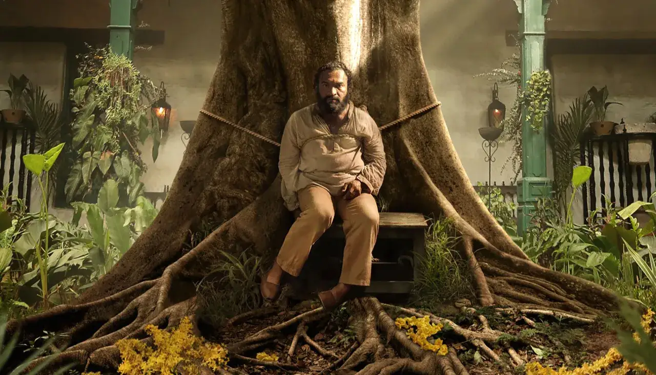 Mężczyzna siedzi przy korzeniach gigantycznego drzewa, otoczony bujną roślinnością. Scena z serialu 