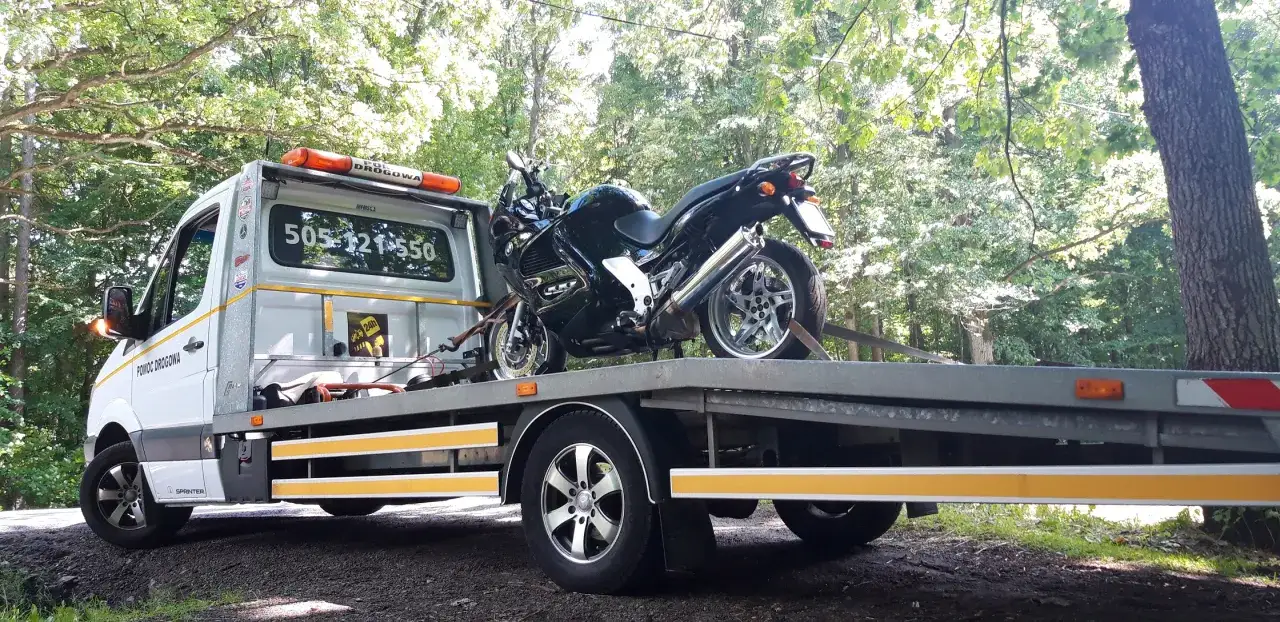Pomoc drogowa holuje motocykl. Na platformie lawety stoi czarny motocykl, zabezpieczony pasami.