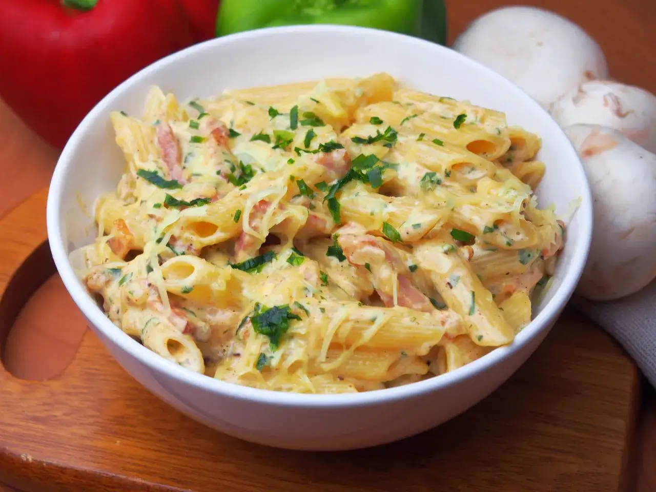 Pyszny makaron z boczkiem w kremowym sosie, posypany świeżą natką pietruszki. Obok leżą pieczarki i papryka.