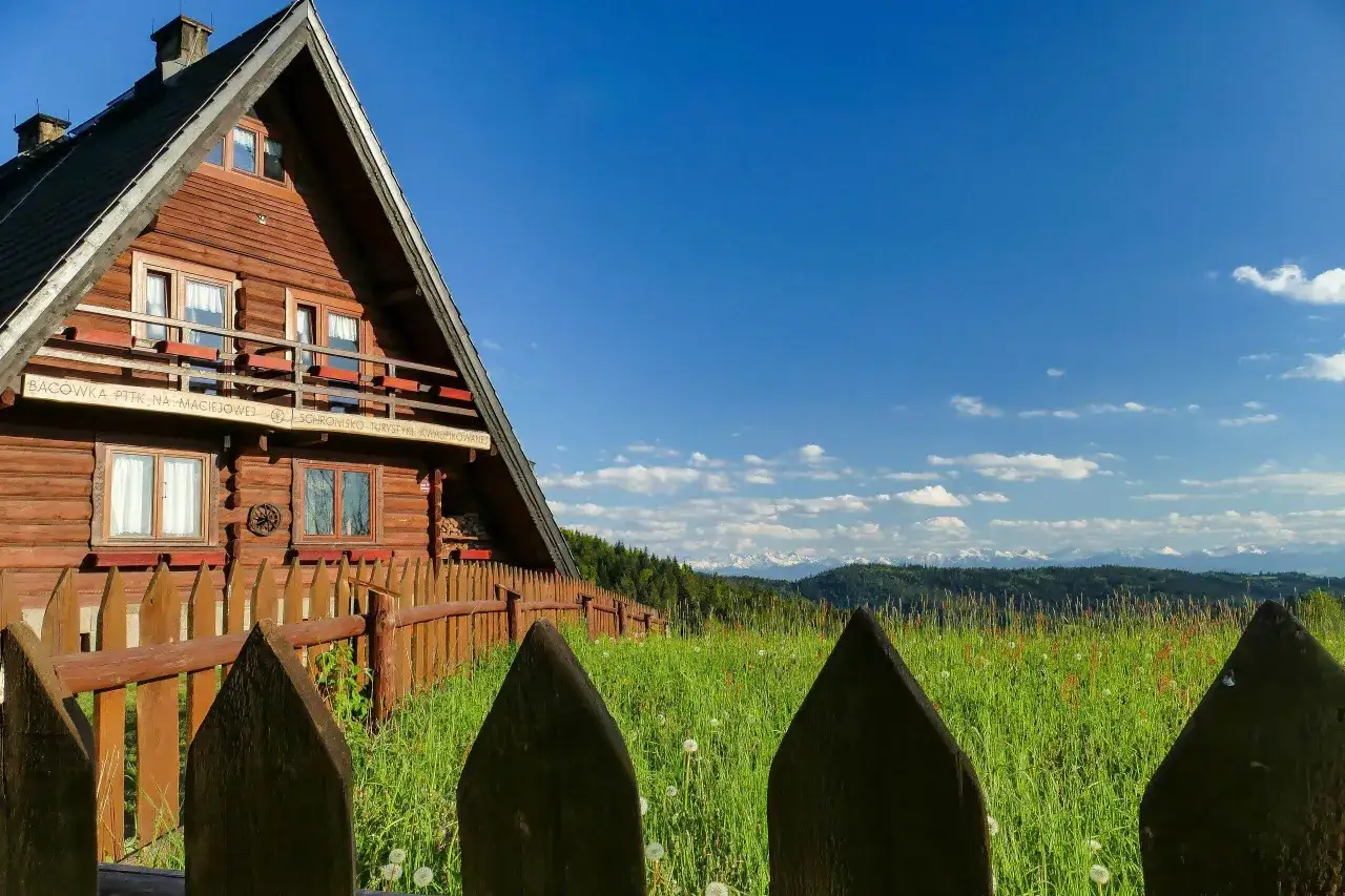 Drewniana bac&oacute;wka na Maciejowej z widokiem na g&oacute;ry. Otoczona zieloną łąką i drewnianym płotem.