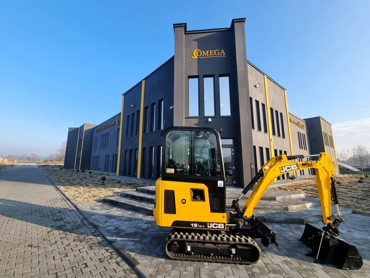 Żółta koparko-ładowarka JCB 19C-1 czeka na rozpoczęcie pracy przed nowoczesnym budynkiem OMEGA.