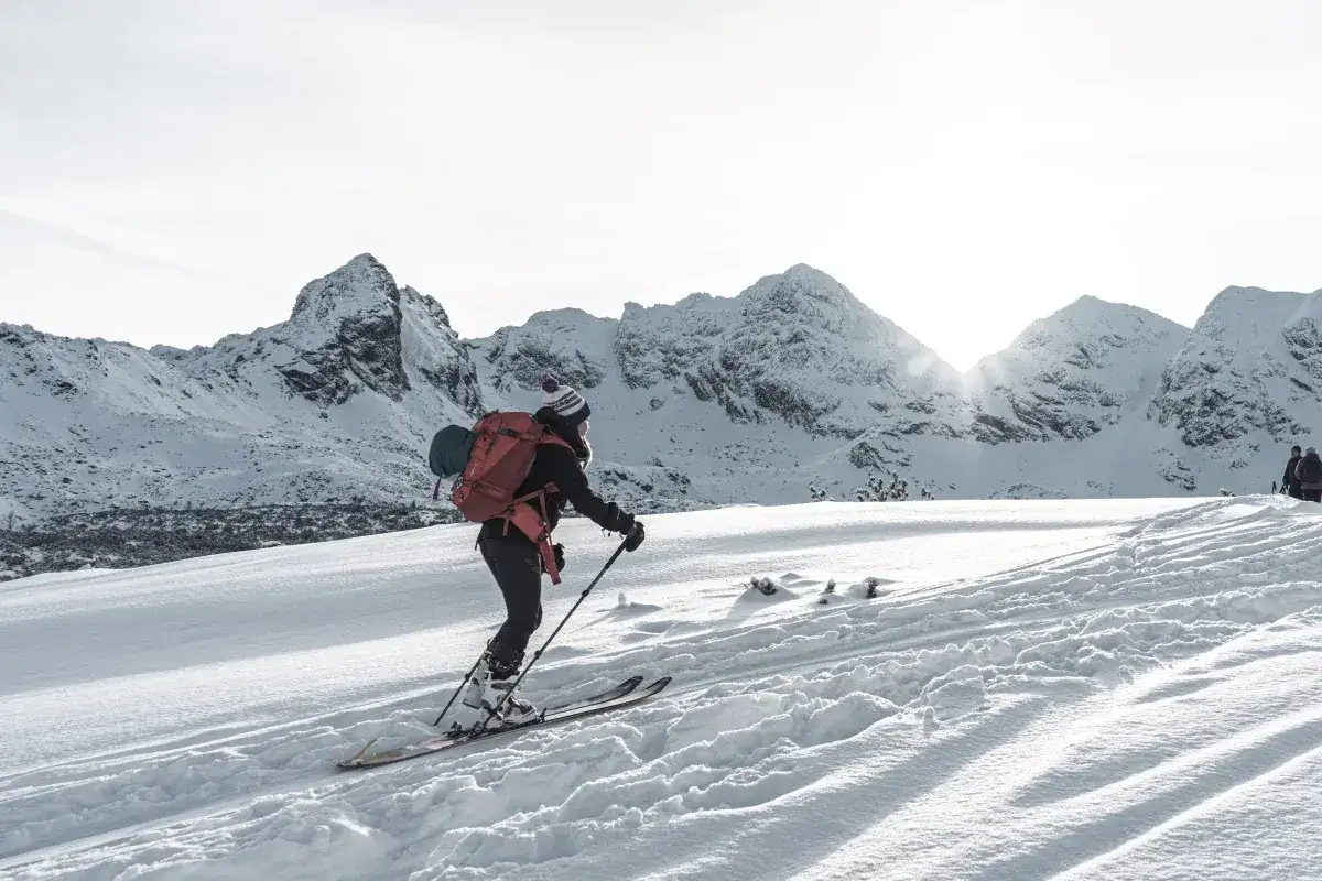 Osoba na nartach z plecakiem przemierza ośnieżone szczyty. Słońce przebija się przez chmury, oświetlając majestatyczne g&oacute;ry. To piękny dzień na ski tury.