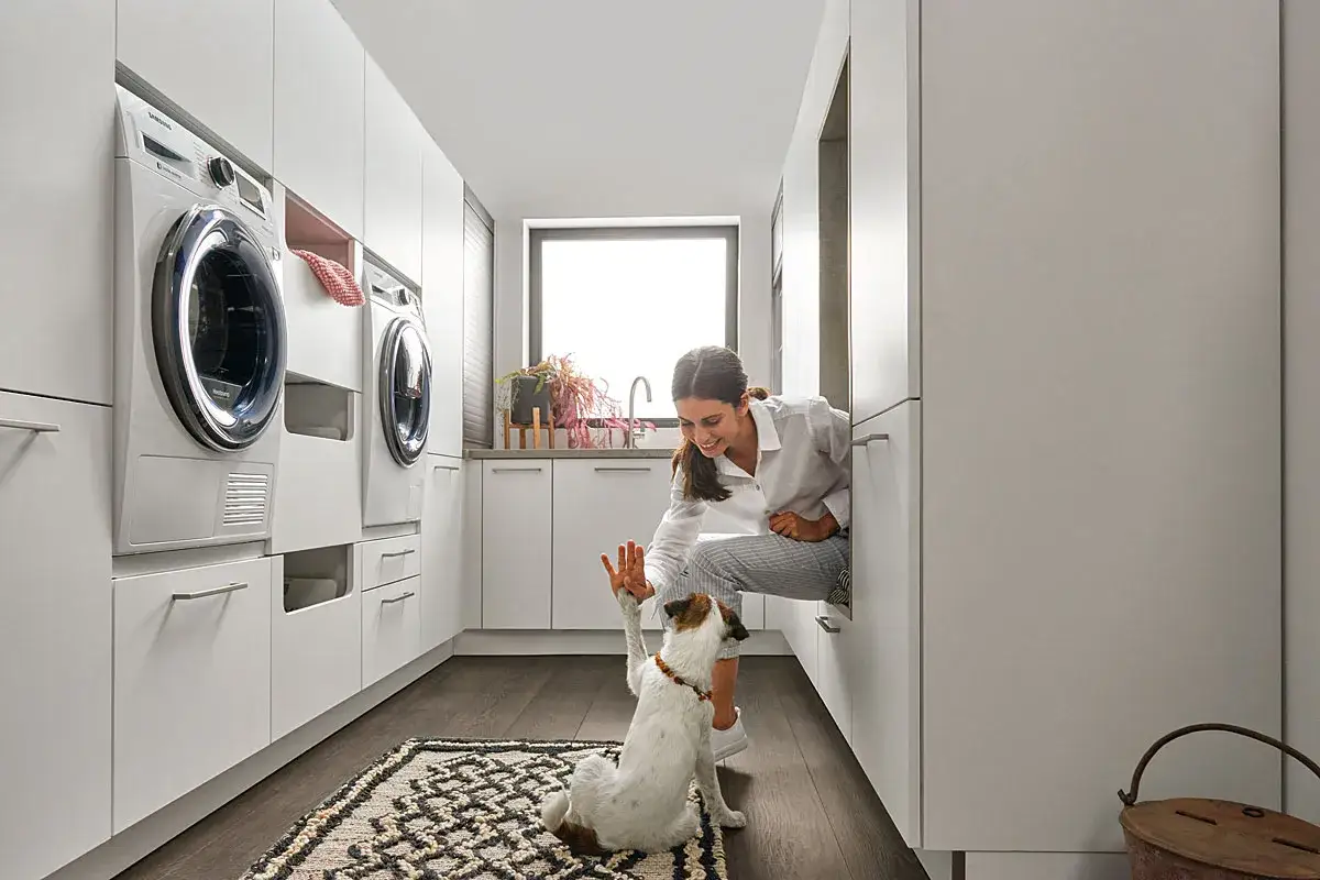 Uśmiechnięta kobieta bawi się z psem w nowoczesnej pralni w domu. W tle pralki i suszarki.