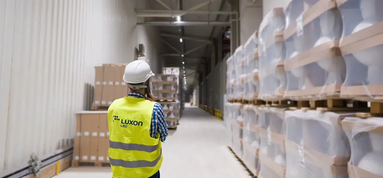 Pracownik w kamizelce odblaskowej Luxon LED wykonuje audyt oświetlenia w magazynie pełnym towar&oacute;w.