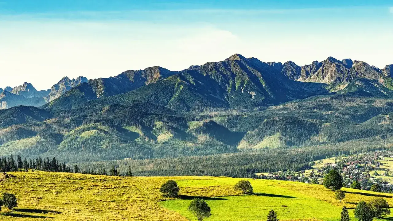 Widok na Tatry z Białki Tatrzańskiej. Zielone łąki i lasy schodzą ku malowniczej wsi, a w tle wznoszą się majestatyczne szczyty gór.