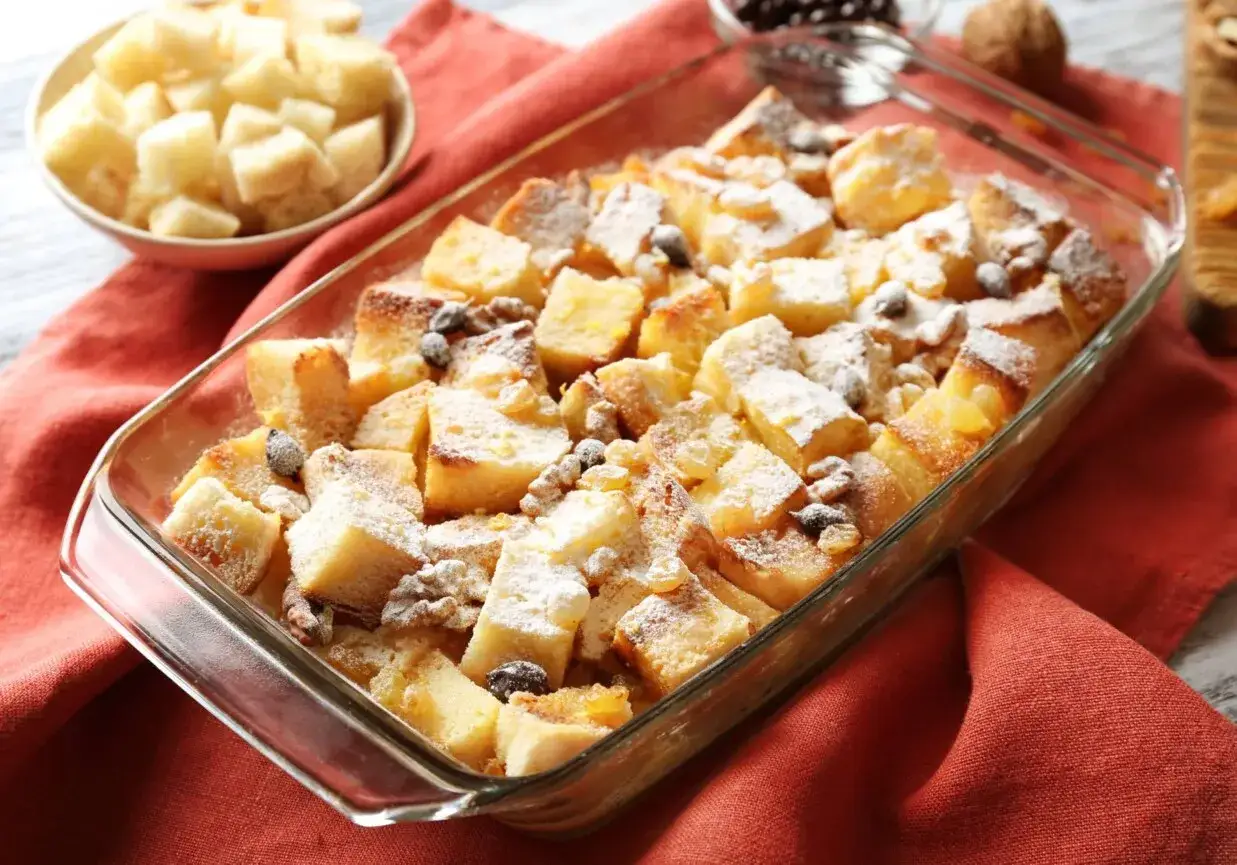 Pudding chlebowy w naczyniu, posypany cukrem pudrem, rodzynkami i orzechami. Obok miseczka z pokrojonym chlebem.