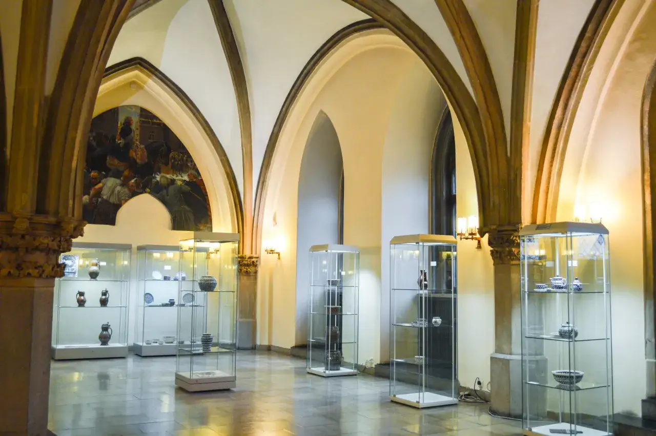 Wnętrze muzeum mieszczaństwa Wrocław. W gablotach eksponaty ceramiczne i metalowe, pod łukami malowidło.