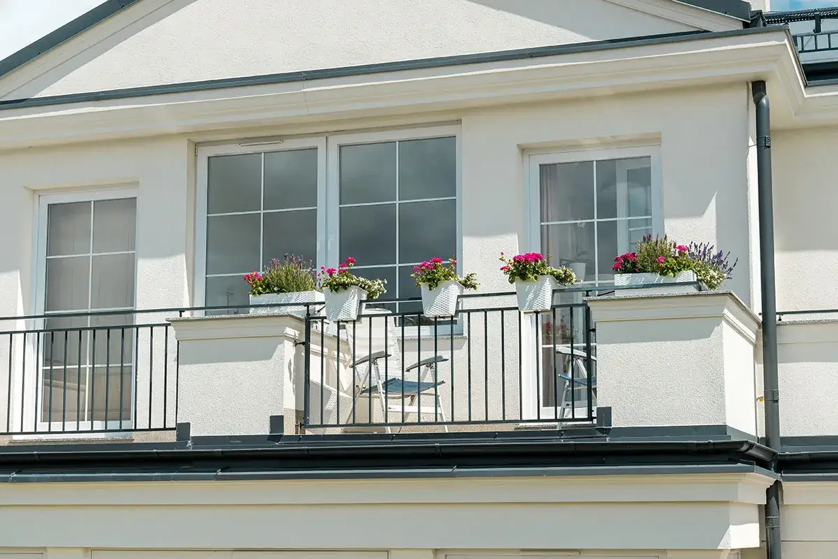 Białe okna pięknie komponują się z jasną, tynkowaną elewacją budynku. Balkon ozdobiony kwiatami dodaje uroku.