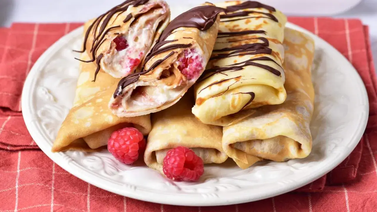 Rolowane naleśniki z malinami i kremowym serkiem mascarpone, polane czekoladą. Pyszny deser na talerzu.