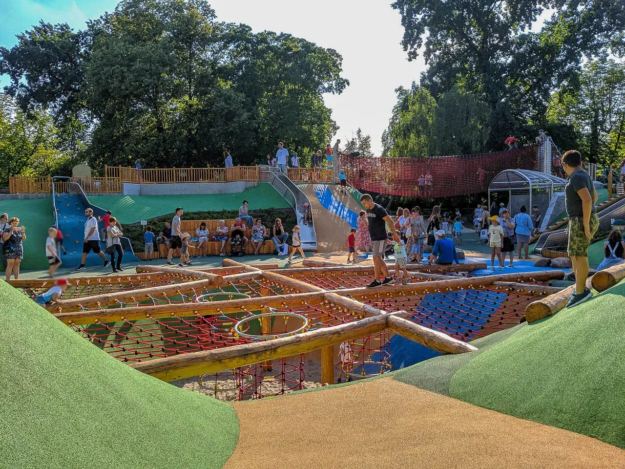 Tętniący życiem plac zabaw w Parku Ujazdowskim, pełen dzieci i dorosłych korzystających z licznych atrakcji, zjeżdżalni i sieciowych konstrukcji.