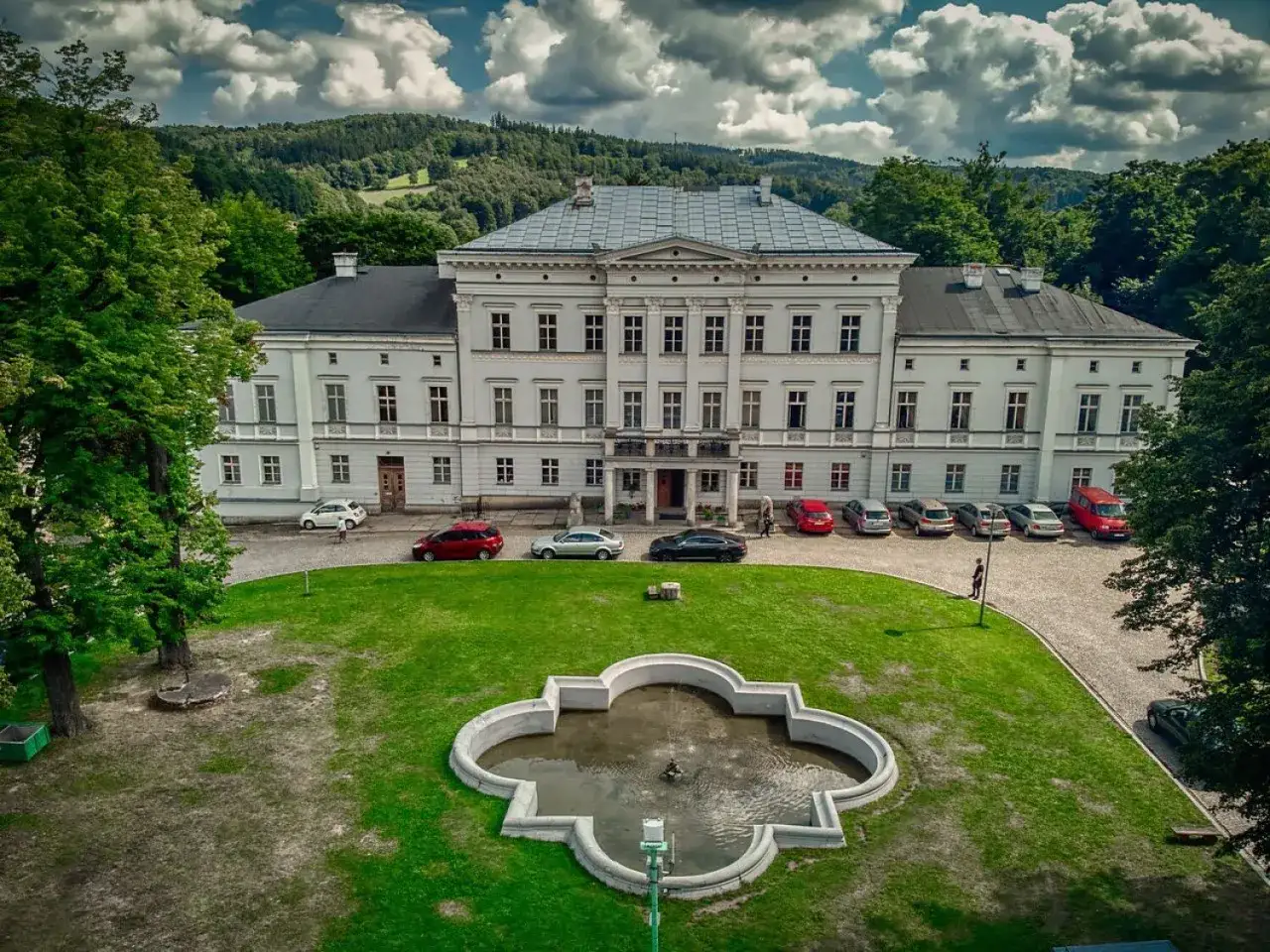 Pałac Jedlinka, biała, okazała budowla z szarym dachem, otoczona zielenią i g&oacute;rami. Przed nim trawnik z fontanną i zaparkowane samochody.