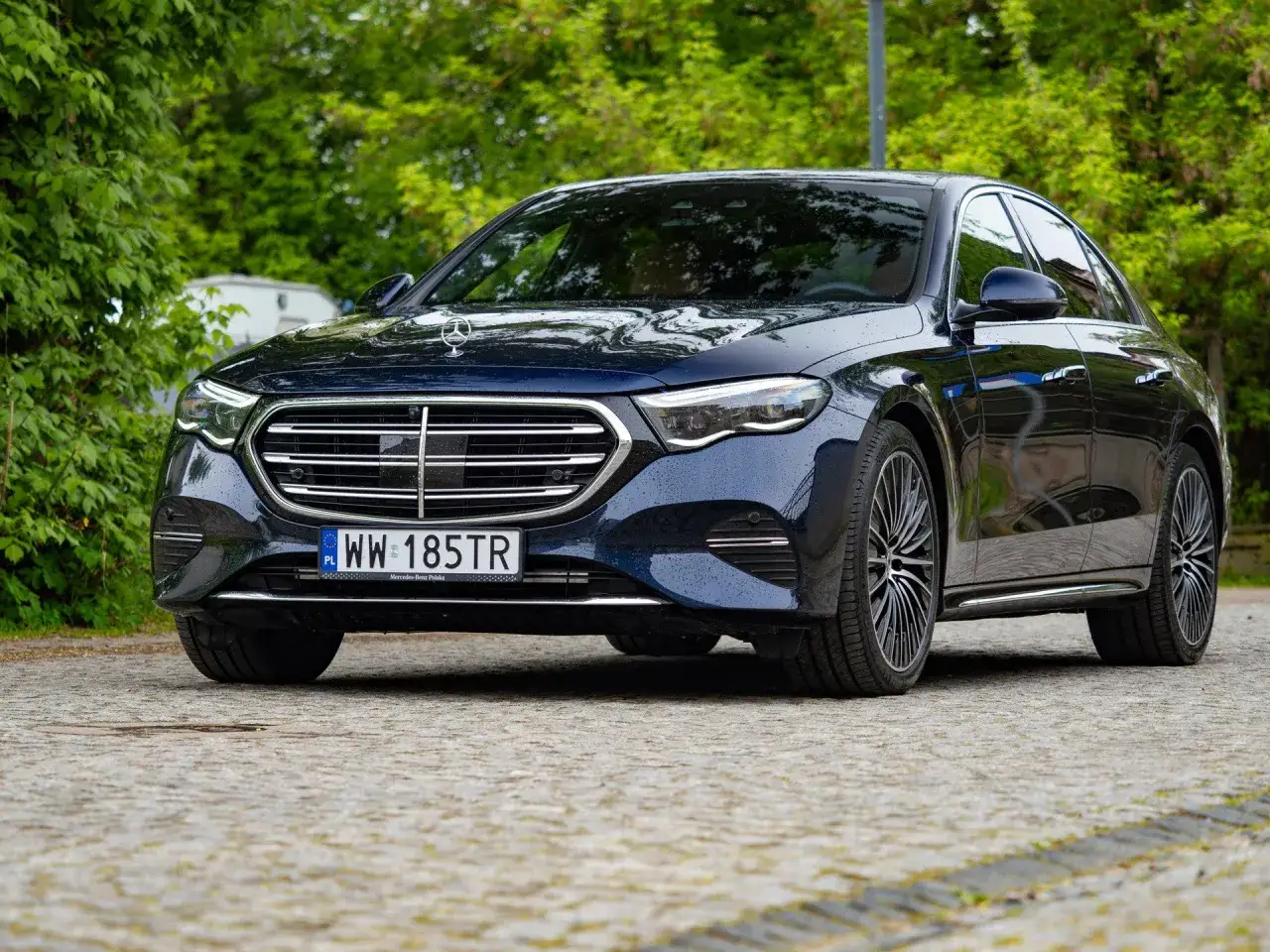 Elegancki, ciemnogranatowy Mercedes-Benz Klasa E z kroplami deszczu na karoserii, zaparkowany na brukowanej nawierzchni wśród zieleni.